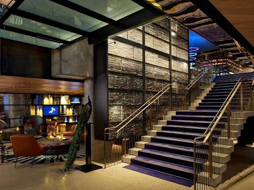QT Melbourne — dramatic interior stairwell with sculptural timber feature wall, brass railings and moody lighting.