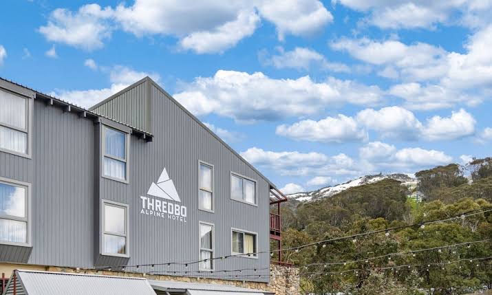 Wide winter photograph of a timber alpine hotel at blue hour — snow-laden gum trees, golden windows, a chairlift disappearing into falling snow, warm smoke curling from a stone chimney.